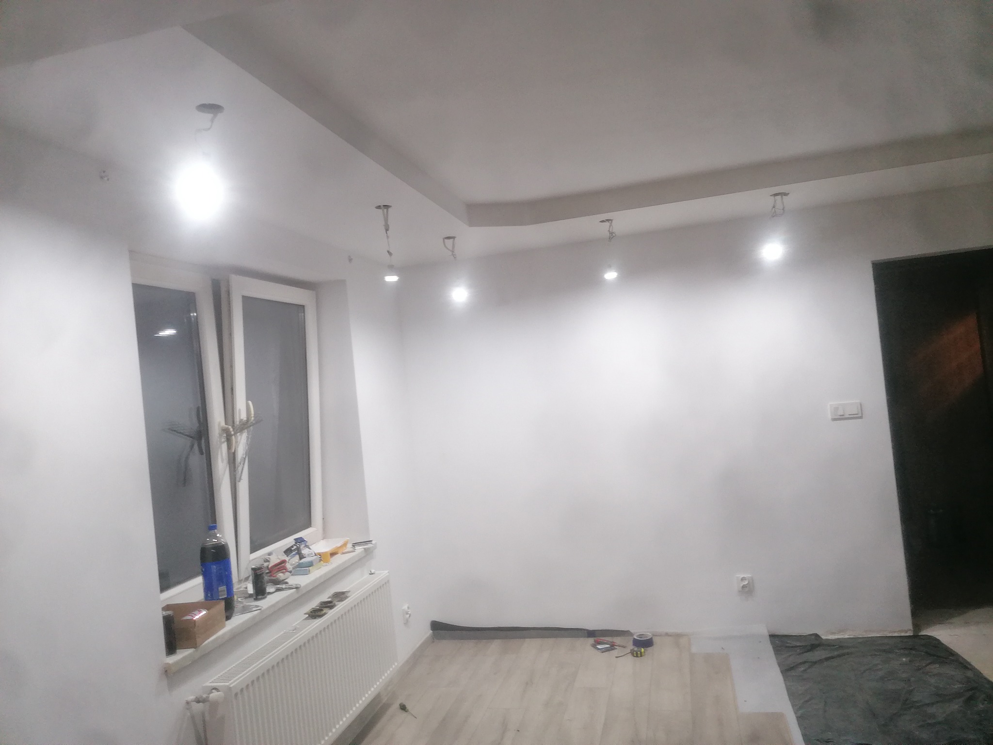Pokój w trakcie prac wykończeniowych: częściowo ułożone panele podłogowe, świeżo pomalowane ściany na biało, widoczne narzędzia i akcesoria malarskie na parapecie, surowe oświetlenie sufitowe.