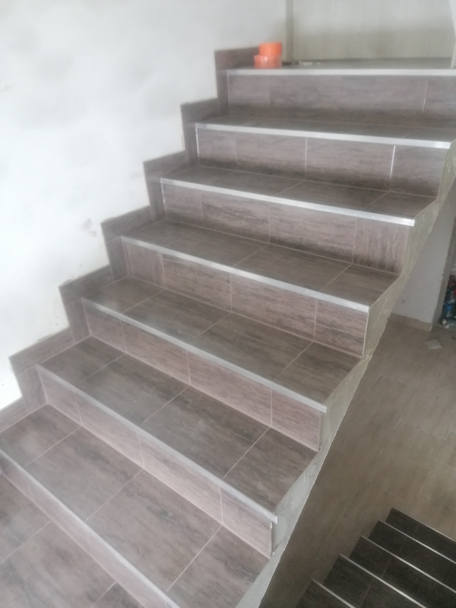 Wykończone schody wewnętrzne wyłożone brązowymi płytkami z aluminiowymi listwami krawędziowymi, widoczne niedociągnięcia w wykończeniu ścian i podłogi.