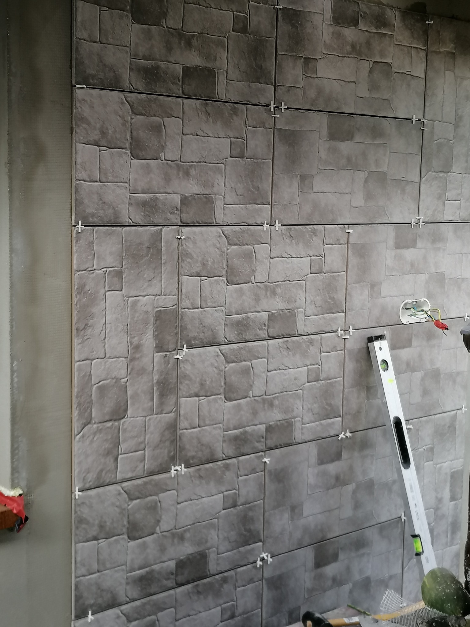 Ściana w trakcie wykańczania z szarymi płytkami imitującymi kamień, widoczne krzyżyki dystansowe, poziomica i wystające kable elektryczne.
