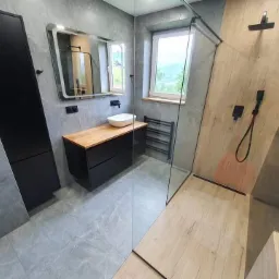 Nowoczesna łazienka z szarymi płytkami imitującymi kamień, drewnianym blatem pod umywalką nablatową, czarnymi szafkami, kabiną prysznicową typu walk-in z drewnopodobnymi panelami na ścianie...