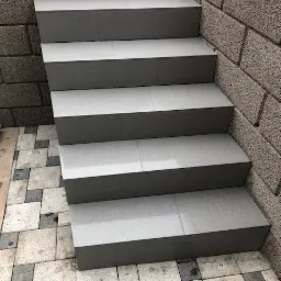 Schody zewnętrzne wyłożone szarą, matową glazurą, z widocznymi fugami, przylegające do ściany z szarego bloczka betonowego, ułożone na kostce brukowej w odcieniach szarości i beżu.