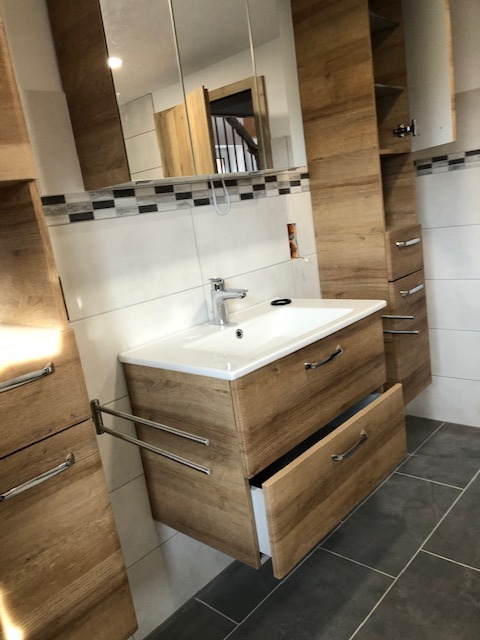 Nowoczesna łazienka z drewnopodobnymi szafkami, umywalką wpuszczaną w blat, lustrem w szafce i szarą podłogą z płytek. Otwarta szuflada pod umywalką ukazuje białe wnętrze.