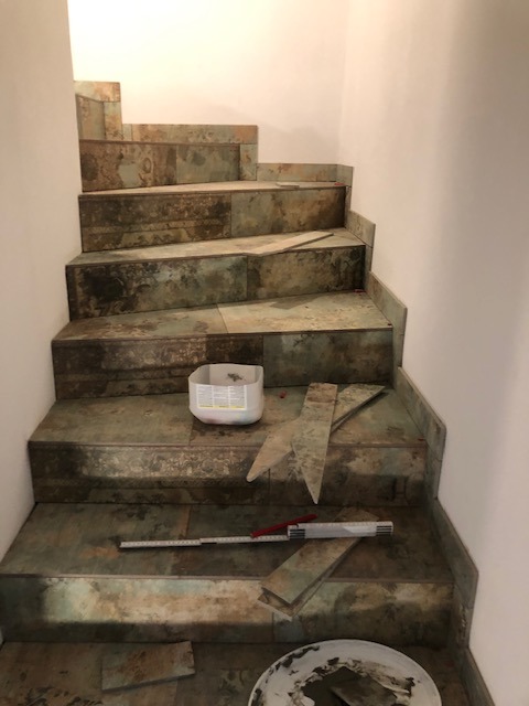 Schody wyłożone płytkami imitującymi kamień, w trakcie prac wykończeniowych, widoczne narzędzia i resztki płytek, białe ściany.