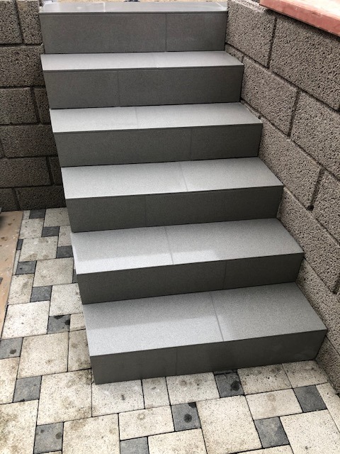 Schody zewnętrzne wyłożone szarą, matową glazurą, z widocznymi fugami, przylegające do ściany z szarego bloczka betonowego, ułożone na kostce brukowej w odcieniach szarości i beżu.