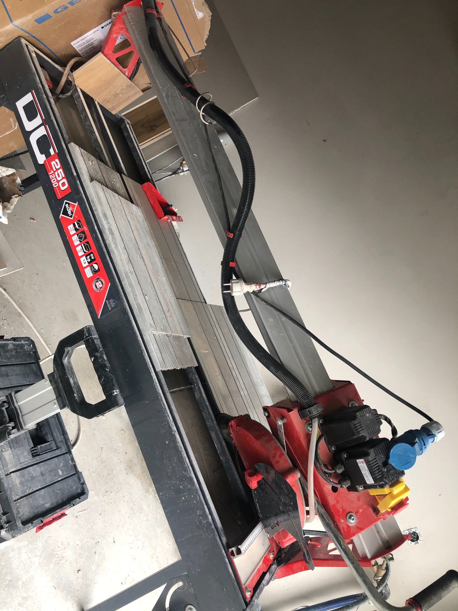 Elektryczna przecinarka do płytek Rubi DC-250 1200 z ułożonymi płytkami gotowa do cięcia, widoczne przewody zasilające i rurka doprowadzająca wodę do chłodzenia tarczy tnącej.