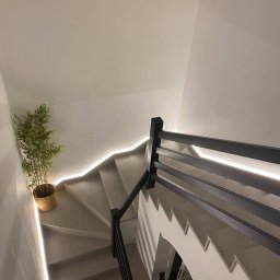 NWCOMPANY SPÓŁKA Z OGRANICZONĄ ODPOWIEDZIALNOŚCIĄ - Betonowe schody spiralne z podświetleniem LED i czarną balustradą, widok z góry. W rogu doniczka z rośliną. Minimalistyczny design.