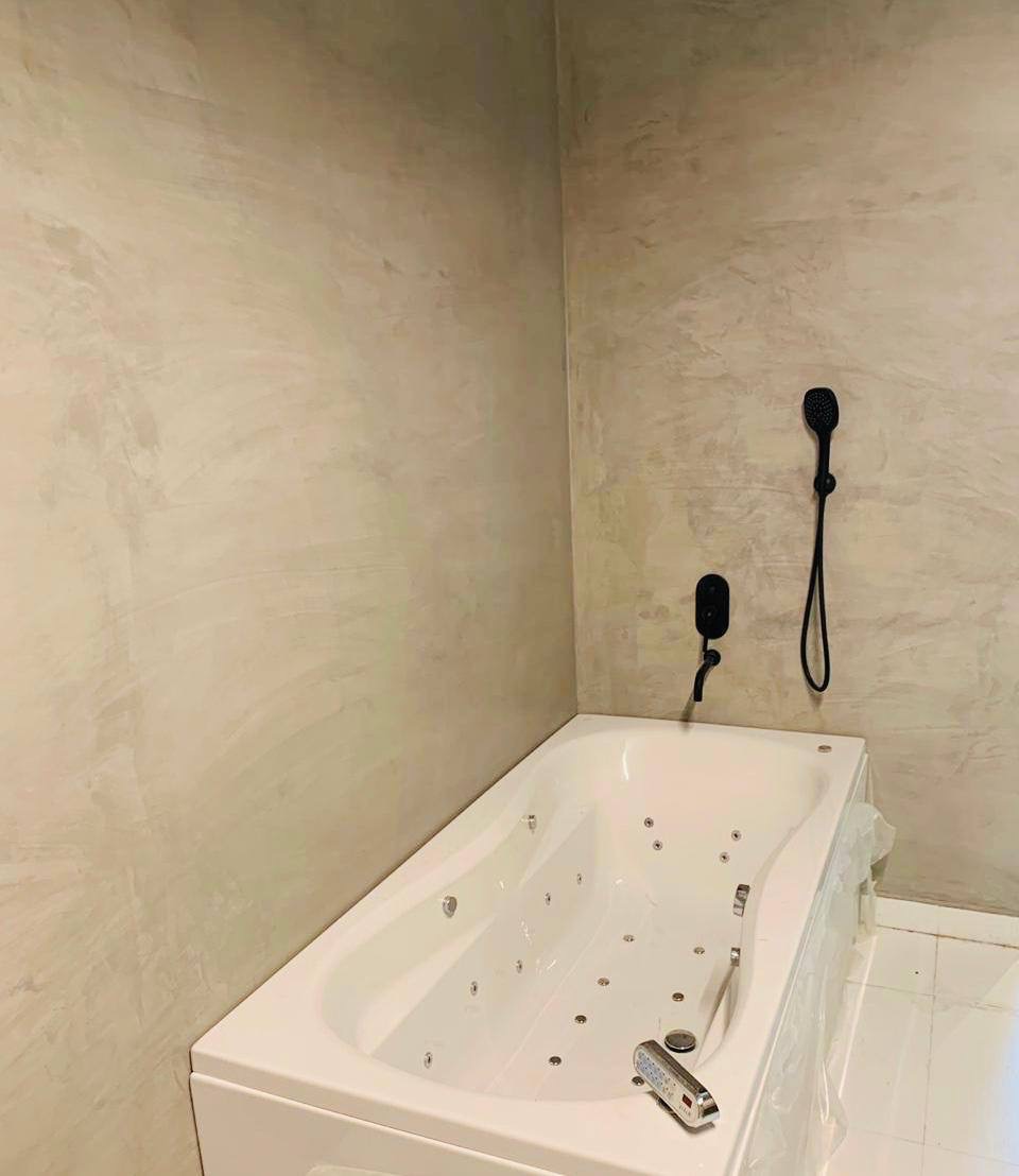 Wykończona łazienka z wanną jacuzzi i ścianami pokrytymi dekoracyjną, beżową gładzią. Widoczna czarna armatura i biała podłoga. Całość w neutralnej tonacji.