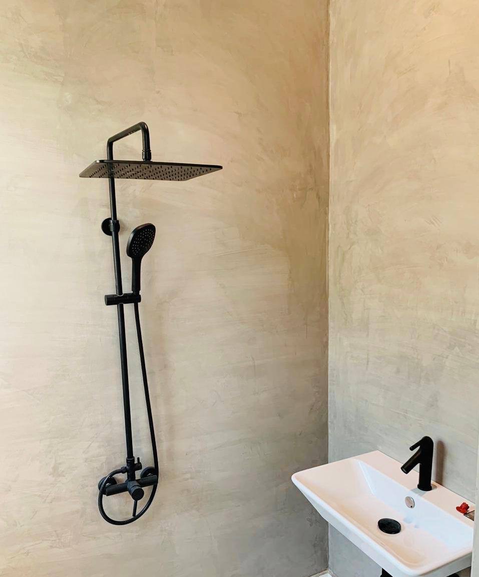 Minimalistyczna łazienka z czarną armaturą: deszczownica i umywalka na tle ściany wykończonej w jasnym, surowym stylu. Widoczne detale wykończenia.