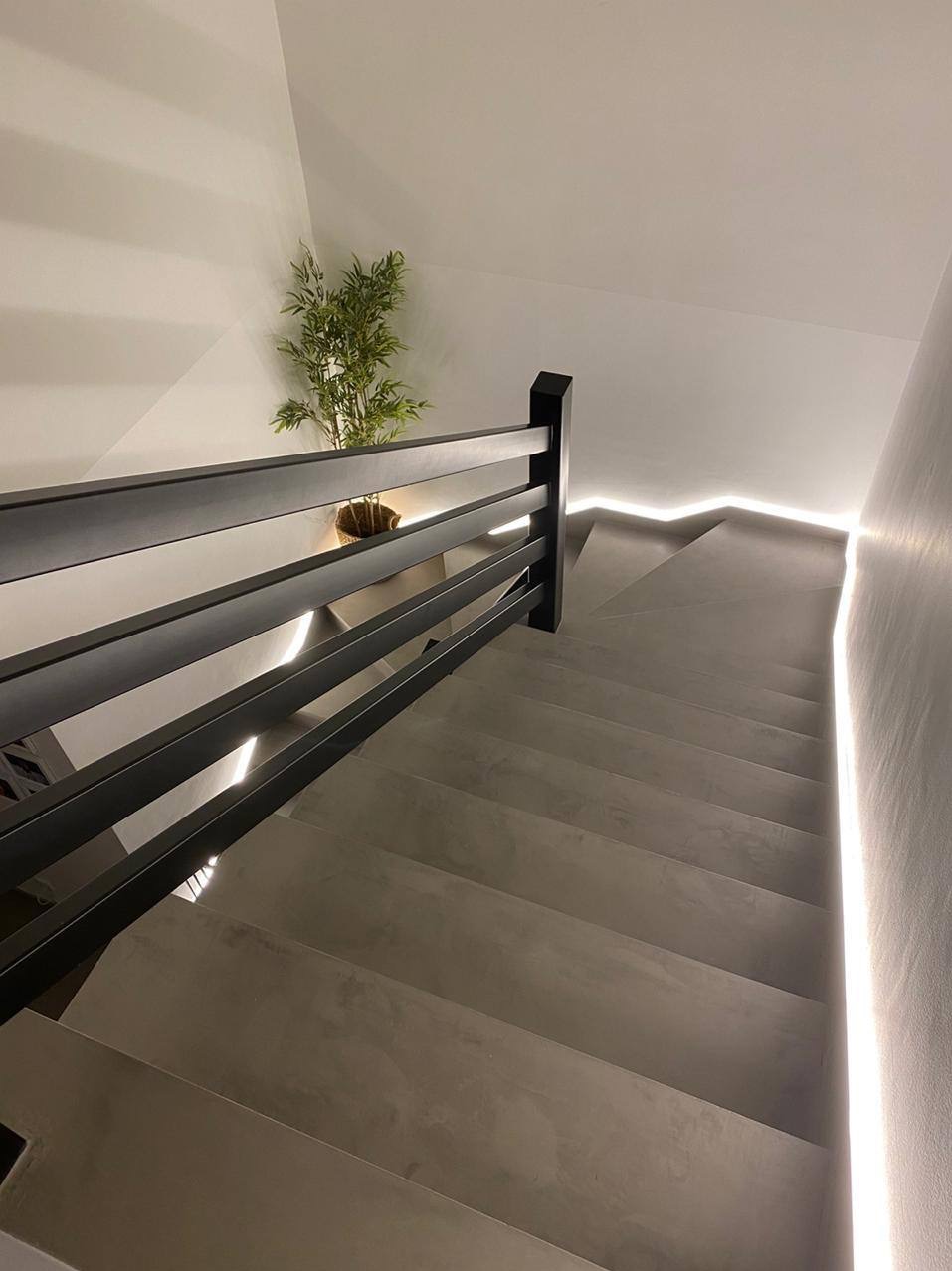 Nowoczesne schody z szarego betonu z czarną balustradą i subtelnym oświetleniem LED wzdłuż ściany. W tle roślina doniczkowa.