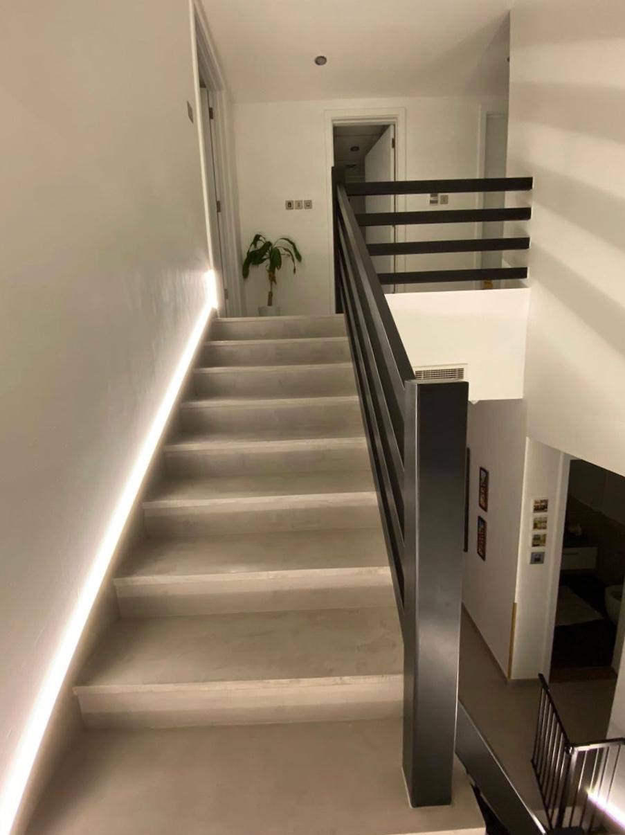 Schody z mikrocementu z podświetleniem LED, czarna balustrada. Minimalistyczny design, jasne wnętrze. Widok z dołu na górę, roślina doniczkowa w tle.