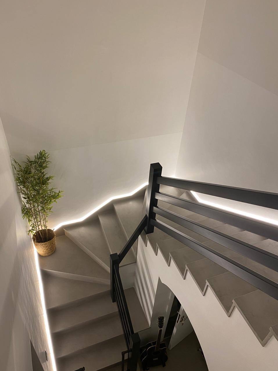 Betonowe schody spiralne z podświetleniem LED i czarną balustradą, widok z góry. W rogu doniczka z rośliną. Minimalistyczny design.