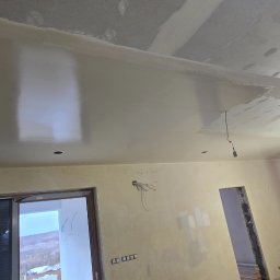DAMAX Remonty i Wykończenia Wnętrz Damian Mruk - Wnętrze w trakcie remontu: częściowo pomalowany sufit z widocznymi śladami szpachlowania, niepomalowane ściany z wystającymi przewodami elektrycznymi, widoczne otwory drzwiowe i okno z widokiem...