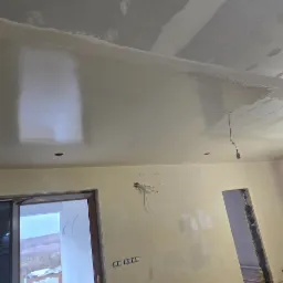 Wnętrze w trakcie remontu: częściowo pomalowany sufit z widocznymi śladami szpachlowania, niepomalowane ściany z wystającymi przewodami elektrycznymi, widoczne otwory drzwiowe i okno z widokiem...