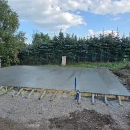 Wylana, świeża płyta betonowa fundamentowa na gruncie, zabezpieczona drewnianym szalunkiem. W tle zieleń drzew i błękitne niebo z chmurami.