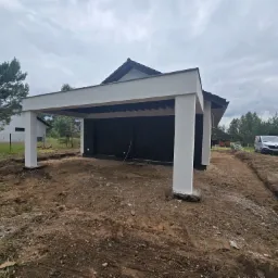 Nowoczesna wiata garażowa z białymi kolumnami i ciemnym dachem, widok z zewnątrz na tle przygotowanego terenu. Widoczny samochód dostawczy w tle.