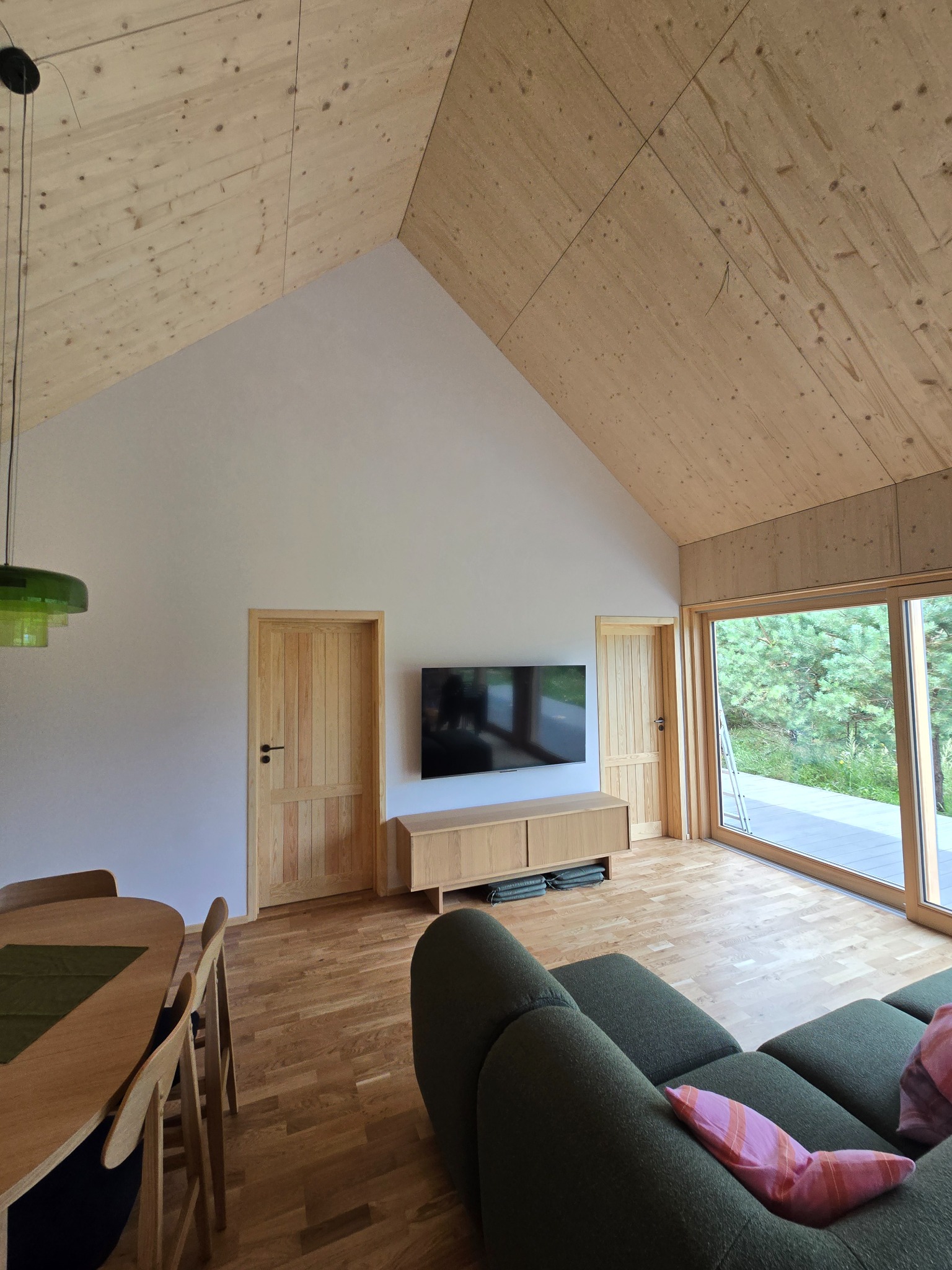 Jasne wnętrze domu ze skośnym, drewnianym sufitem i drewnianą podłogą. Widoczne drzwi z naturalnego drewna i duże okno przesuwne z widokiem na zieleń.