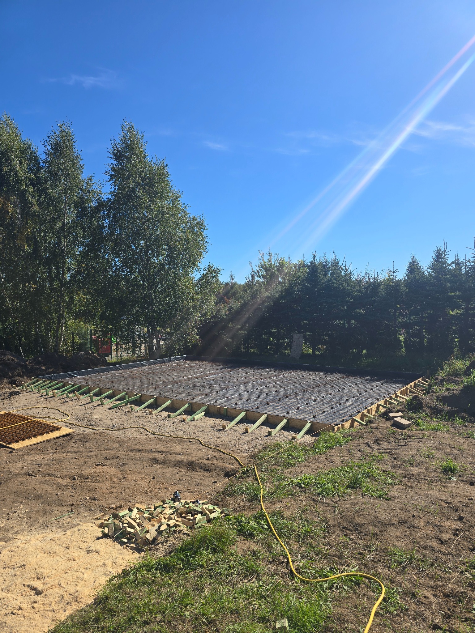 Przygotowanie podłoża pod wylewkę: ułożona folia, siatka zbrojeniowa na legarach. Widoczne drewniane elementy i rury. Tło: drzewa i błękitne niebo z refleksami słońca.