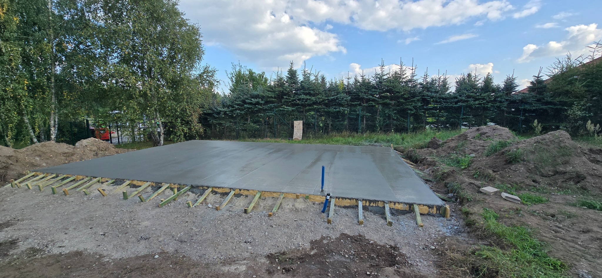 Wylana, świeża płyta betonowa fundamentowa na gruncie, zabezpieczona drewnianym szalunkiem. W tle zieleń drzew i błękitne niebo z chmurami.