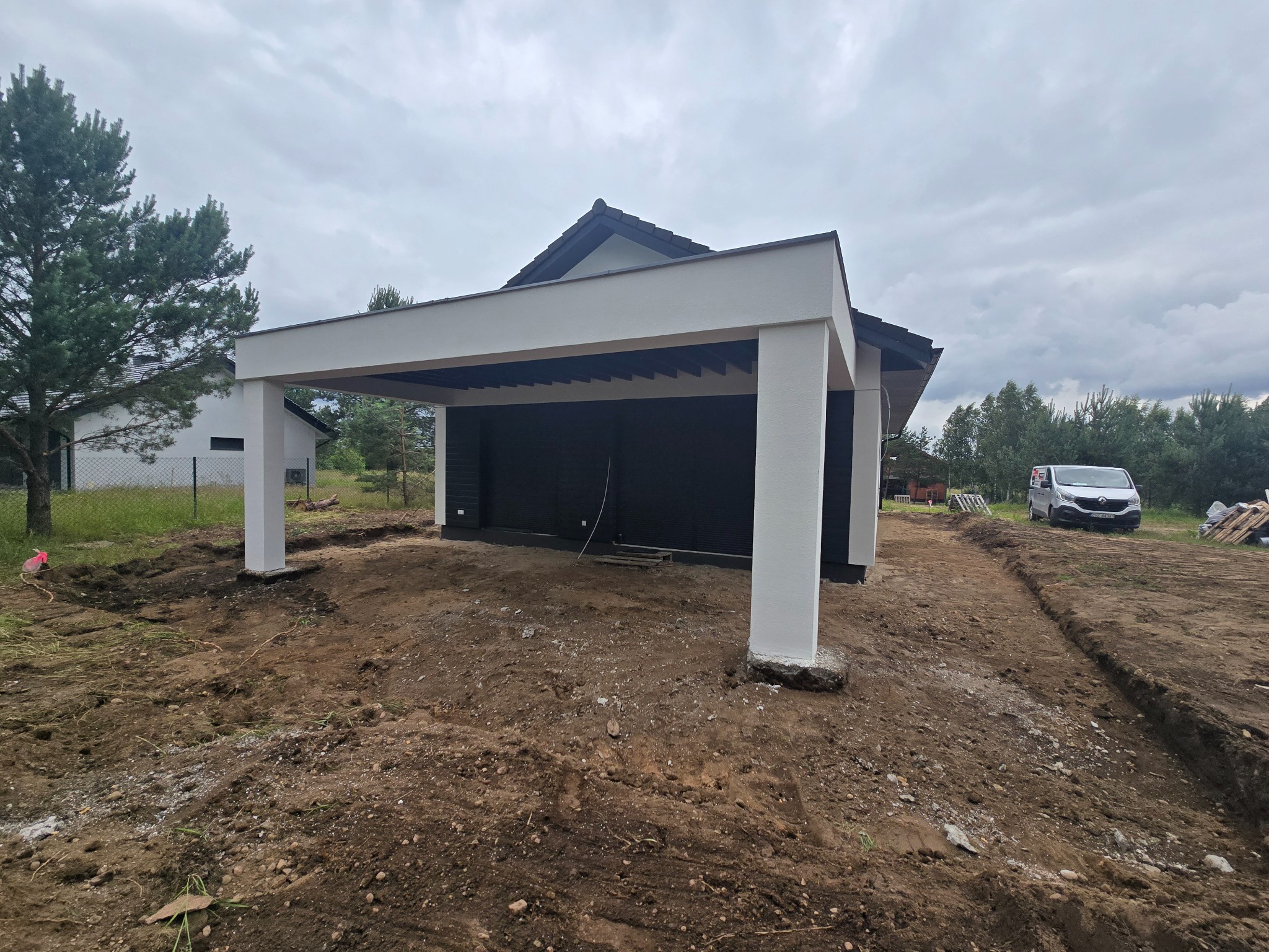 Nowoczesna wiata garażowa z białymi kolumnami i ciemnym dachem, widok z zewnątrz na tle przygotowanego terenu. Widoczny samochód dostawczy w tle.