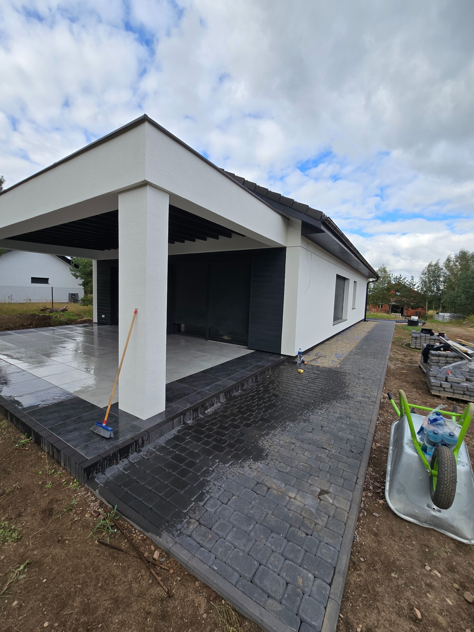 Nowoczesna aranżacja przestrzeni: ciemna kostka brukowa prowadzi do zadaszonego tarasu z szarych płyt. Widoczny wózek i narzędzia sugerują świeżo zakończone prace.