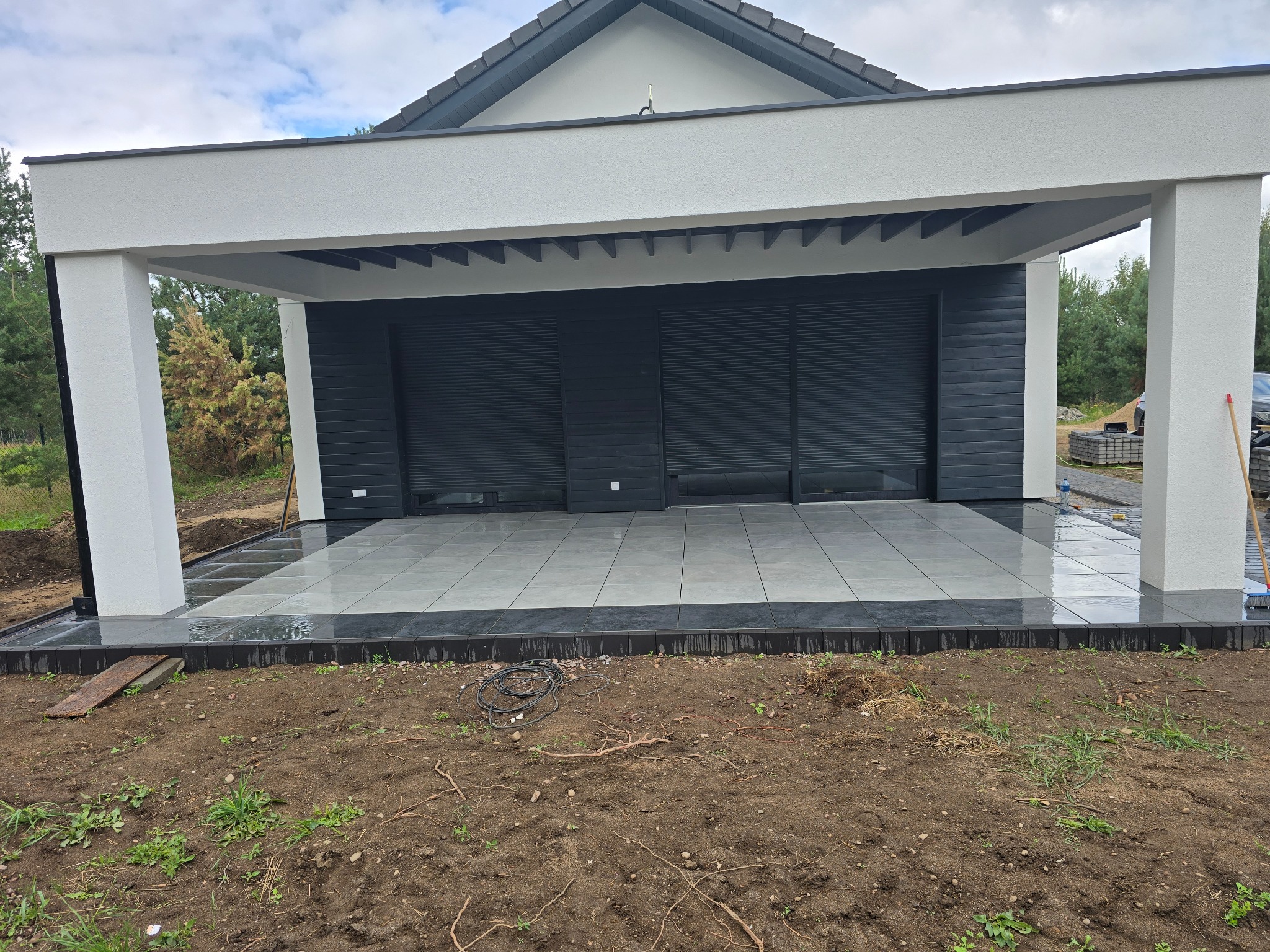 Nowoczesna wiata garażowa z szarymi płytkami i czarnymi roletami, otoczona ziemią. Widoczne detale wykończenia i minimalistyczny design.