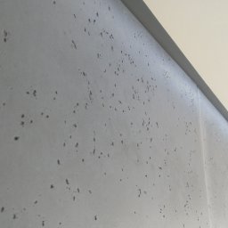 Impuls Energia - Szara ściana z efektem betonu architektonicznego, widoczna z bliska pod kątem, z fragmentem sufitu i zasłony w tle. Minimalistyczny design wnętrza.