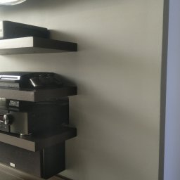 Impuls Energia - Ciemne, minimalistyczne półki wiszące na szarej ścianie, na których stoją sprzęty RTV: konsola, wzmacniacz i głośnik. Widok z dołu, oświetlenie dzienne.