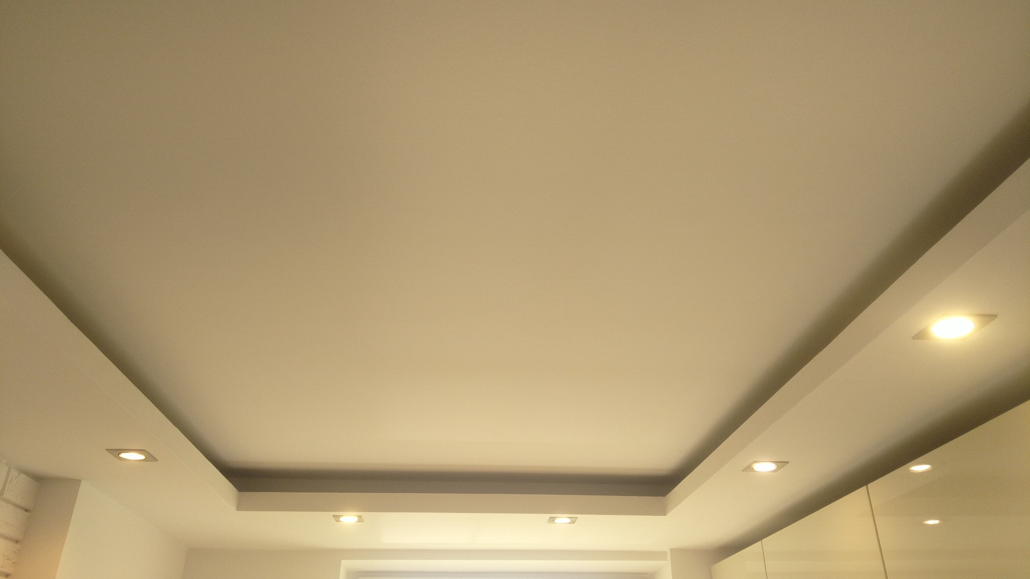 Podwieszany sufit z wbudowanymi, kwadratowymi lampami LED, oświetlający wnętrze. Widoczne fragmenty ścian i mebli w jasnych barwach.