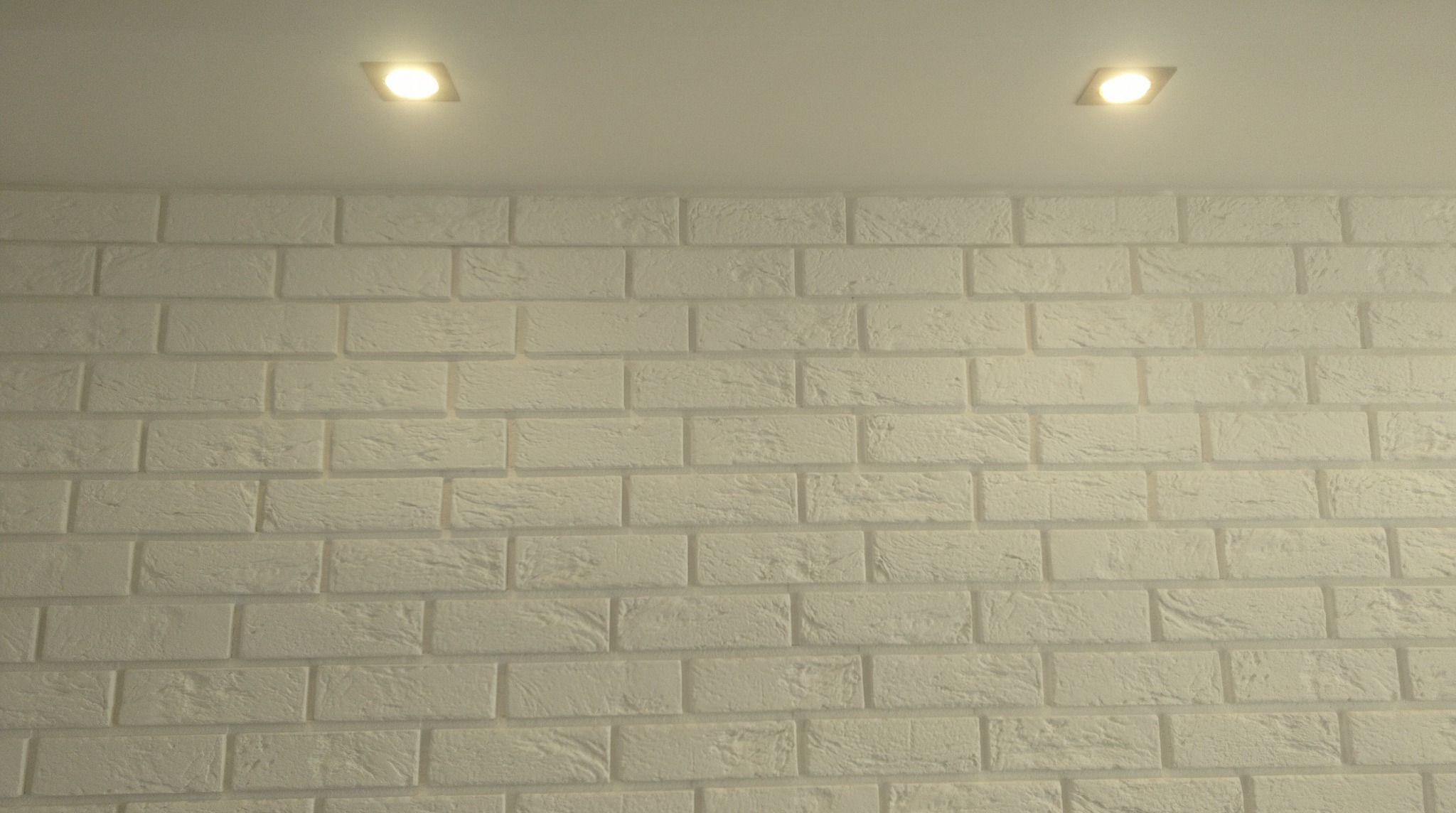 Biała ściana z cegieł dekoracyjnych podświetlona dwoma punktowymi lampami sufitowymi. Tekstura cegły wyraźnie widoczna, ciepłe światło lamp.