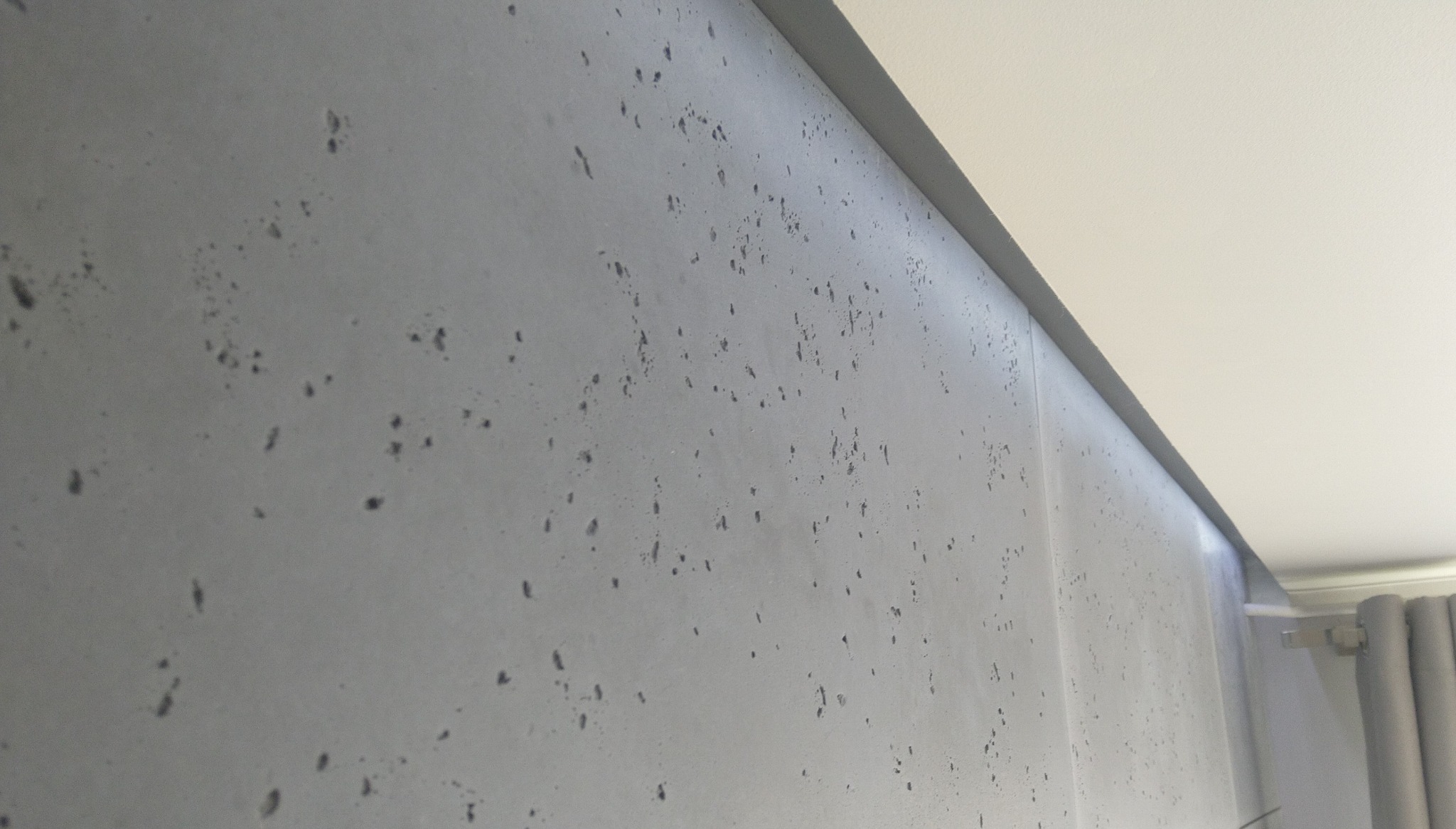 Szara ściana z efektem betonu architektonicznego, widoczna z bliska pod kątem, z fragmentem sufitu i zasłony w tle. Minimalistyczny design wnętrza.
