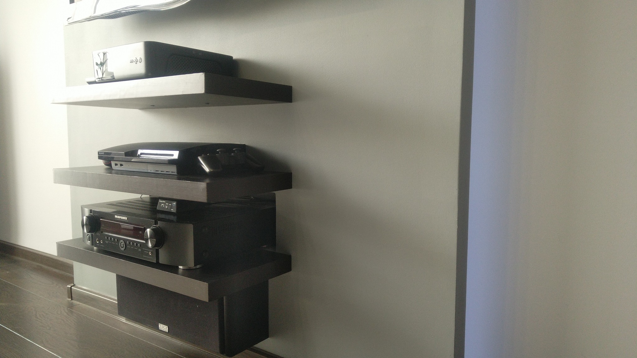 Ciemne, minimalistyczne półki wiszące na szarej ścianie, na których stoją sprzęty RTV: konsola, wzmacniacz i głośnik. Widok z dołu, oświetlenie dzienne.