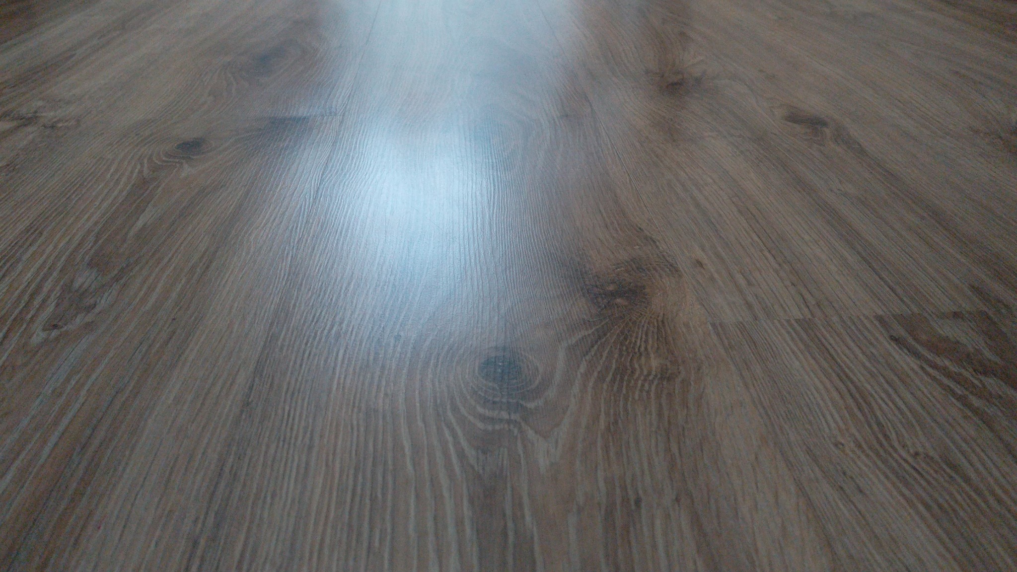 Ułożone panele podłogowe w odcieniach brązu, z widoczną strukturą drewna i delikatnym połyskiem, sfotografowane pod kątem, ukazujące perspektywę podłogi.