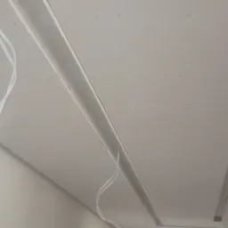 Sufit podwieszany z płyt kartonowo-gipsowych w trakcie montażu, widoczne profile metalowe, przewody elektryczne i łączenia płyt. W tle surowa ściana z widocznymi otworami okiennymi.