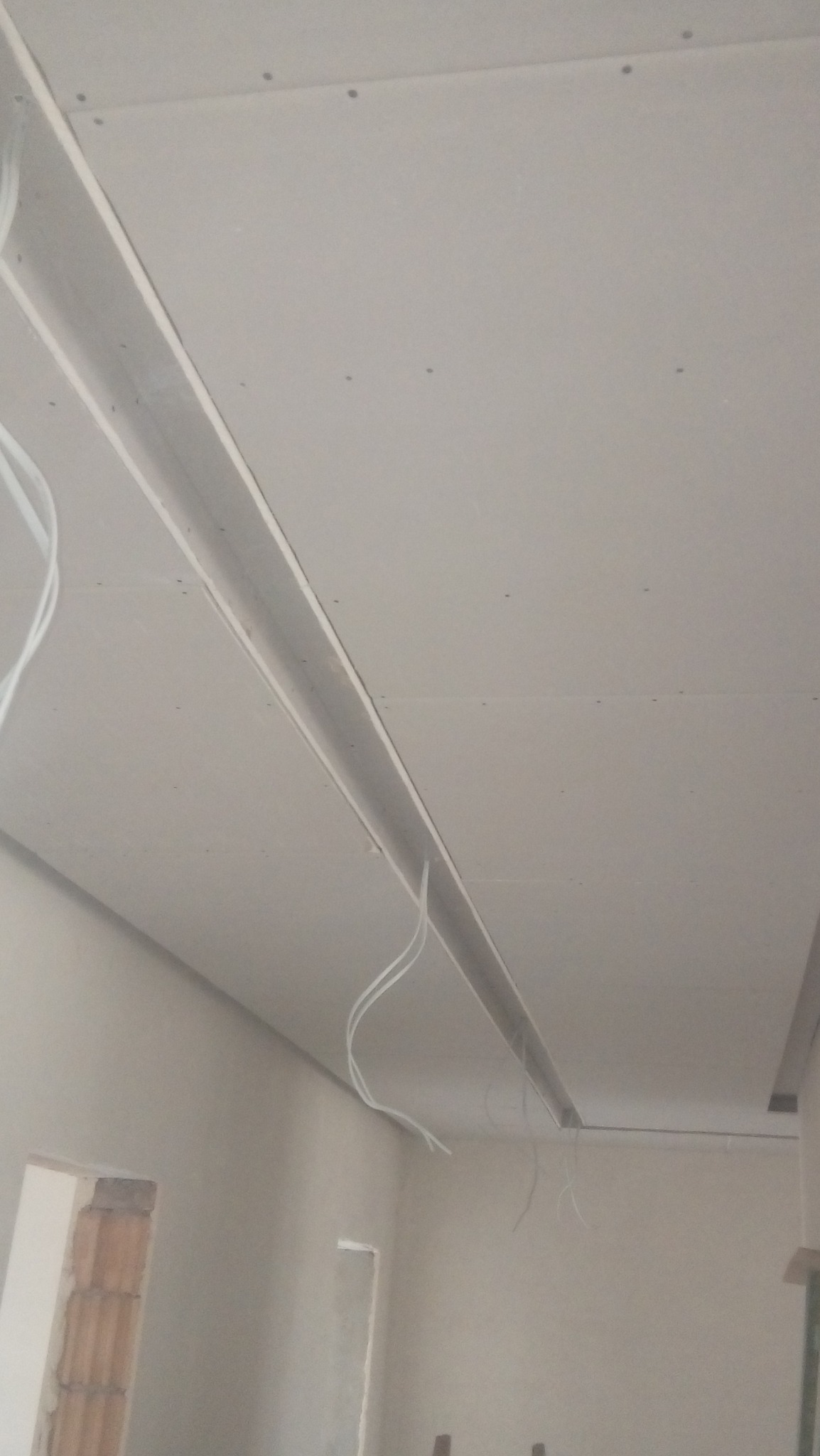 Sufit podwieszany z płyt kartonowo-gipsowych w trakcie montażu, widoczne profile metalowe, przewody elektryczne i łączenia płyt. W tle surowa ściana z widocznymi otworami okiennymi.