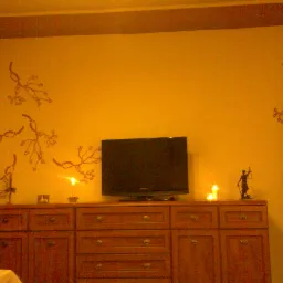 Żółta ściana z ręcznie malowanymi gałązkami, brązowa komoda z telewizorem i świecami, widoczna figurka Temidy, ciepłe oświetlenie.