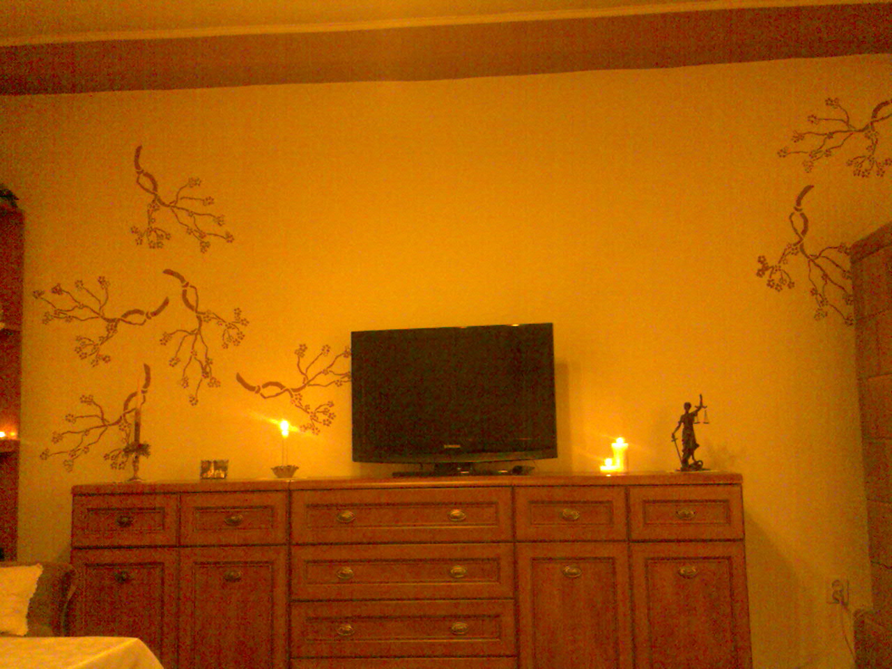 Żółta ściana z ręcznie malowanymi gałązkami, brązowa komoda z telewizorem i świecami, widoczna figurka Temidy, ciepłe oświetlenie.