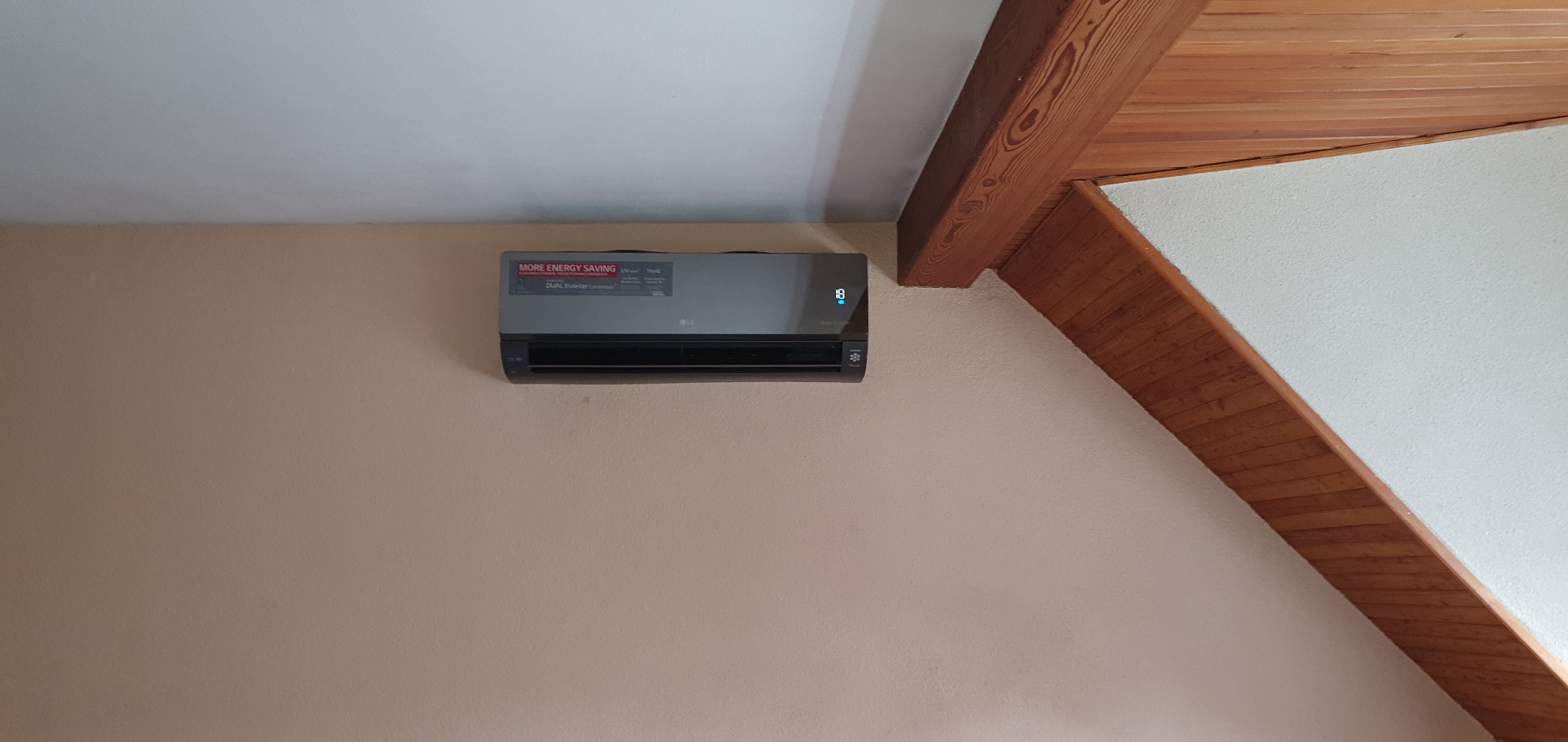 Szara klimatyzacja LG zamontowana na beżowej ścianie w pomieszczeniu z drewnianą belką stropową.
