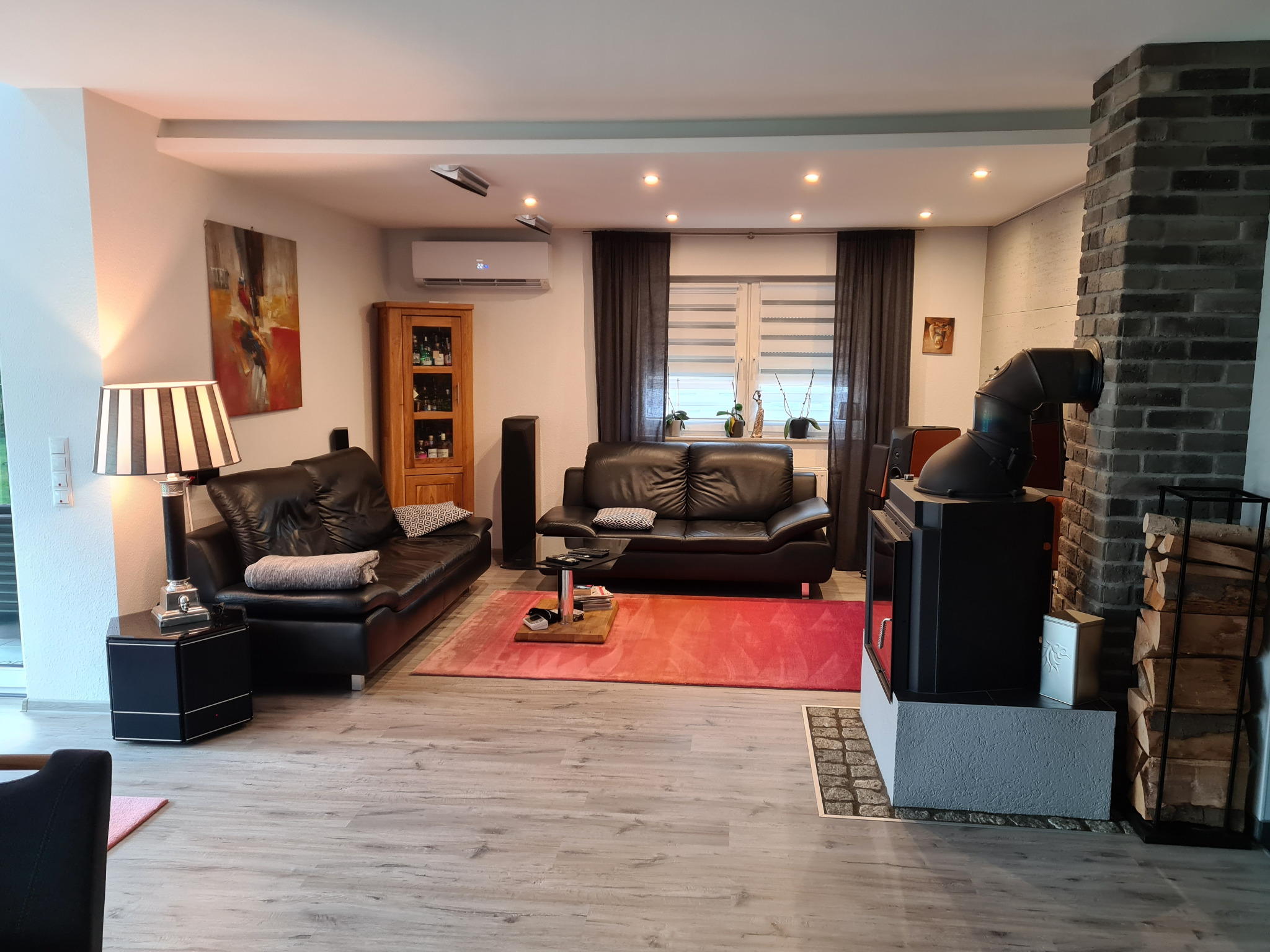Nowoczesny salon z klimatyzatorem ściennym, kominkiem z zapasem drewna, dwiema skórzanymi sofami i czerwonym dywanem.