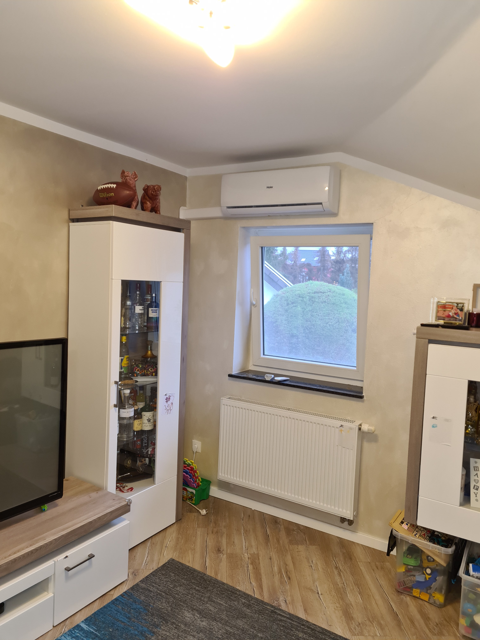 Biały klimatyzator ścienny marki Haier zamontowany w salonie nad oknem, widok na zadbany ogród, po lewej stronie regał z alkoholem, dekoracje w postaci piłki do futbolu amerykańskiego i figurek...