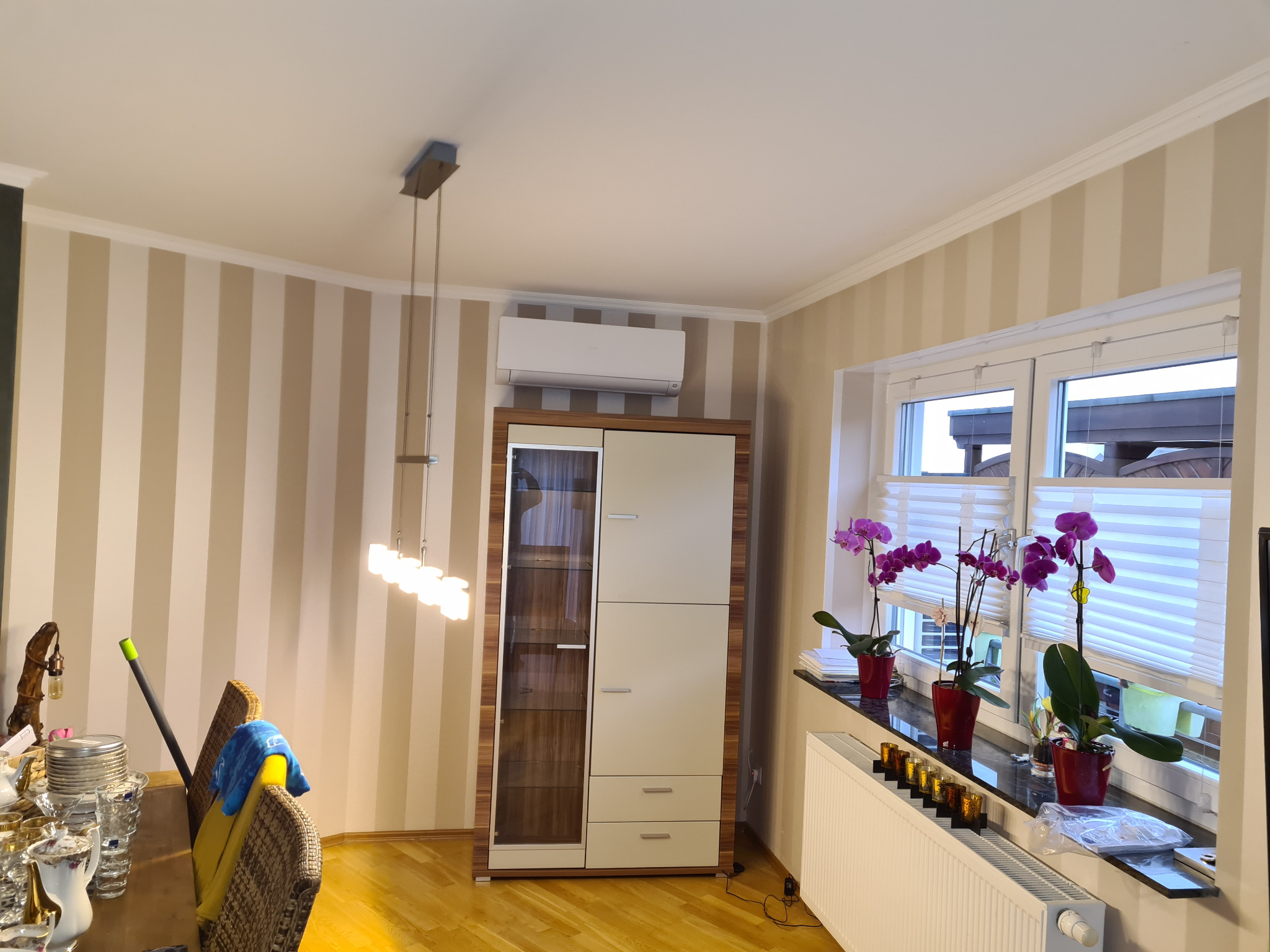 Wnętrze pokoju z zainstalowaną jednostką klimatyzacyjną nad wysokim meblem, widoczne kwiaty w donicach na parapecie okiennym.