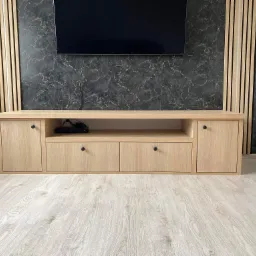 Jasna, drewniana szafka RTV z dwoma szafkami i dwiema szufladami, z czarnymi gałkami, na tle ściany z imitacją czarnego marmuru oraz drewnianych lameli, pod wiszącym telewizorem.