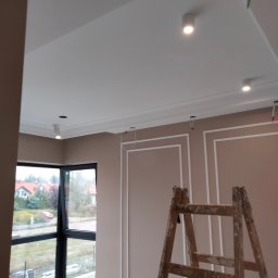 ProfeSSionallToollSsWwaOkolice - Wnętrze w Warszawie po remoncie, z pomalowanymi ścianami w odcieniach beżu, sztukaterią, białym sufitem z oświetleniem punktowym i drewnianą drabiną. Widok na zewnątrz przez okno.