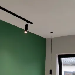 Ściana pomalowana na zielono z zamontowanym czarnym systemem szynowym oświetlenia punktowego oraz pojedyncza lampa wisząca na tle okna.