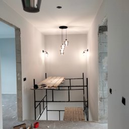 ProfeSSionallToollSsWwaOkolice - Wnętrze w trakcie remontu: białe ściany, surowy betonowy podkład, rusztowanie z drewnianym pomostem, narzędzia i akcesoria budowlane rozłożone na podłodze, wiszące lampy.