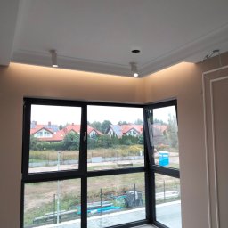 ProfeSSionallToollSsWwaOkolice - Wnętrze pokoju z dużym narożnym oknem z czarną ramą, widok na domy z panelami słonecznymi, sufit z oświetleniem ledowym i ozdobną sztukaterią, ściany w kolorze beżowym z dekoracyjnymi listwami.