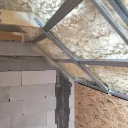 Poddasze w trakcie izolacji pianą natryskową, widoczne metalowe profile konstrukcyjne, ściana z betonu komórkowego i płyta OSB.