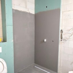 ProfeSSionallToollSsWwaOkolice - Niedokończona łazienka z szarymi ścianami, częściowo pokrytymi gładzią, widoczne płyty gipsowo-kartonowe, białe bloczki, otwarta toaleta i odpływ liniowy w podłodze.