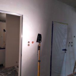 ProfeSSionallToollSsWwaOkolice - Wnętrze w trakcie prac wykończeniowych: ściana po nałożeniu gładzi, zabezpieczone folią drzwi, widoczne puszki elektryczne i lampa robocza na statywie.