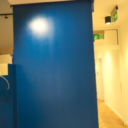 ProfeSSionallToollSsWwaOkolice - Świeżo pomalowana na niebiesko ściana z taśmą malarską zabezpieczającą krawędzie, widoczny fragment podłogi z parkietem w jodełkę i oświetlenie sufitowe z oznaczeniem drogi ewakuacyjnej.