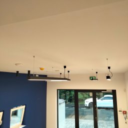 ProfeSSionallToollSsWwaOkolice - Wnętrze w trakcie prac wykończeniowych: pomalowana na granatowo ściana z oknami, biały sufit z nowoczesnym oświetleniem, podłoga częściowo zakryta folią, widoczne butelki wody i narzędzia.